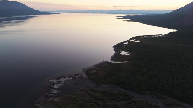 Evening light traces a sinuous coastline where a river meets Lake Lama. The drone glides toward a distant headland through clear air and silence. Putorana Plateau remains wild and inviting