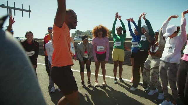 A group of joyful runners with race bibs celebrates success outdoors on a road. They cheer and raise their arms, expressing happiness and teamwork after completing a physical challenge.