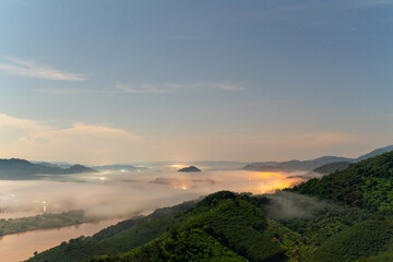 A magical night view from Phu Pha Dak, Nong Khai, Thailand. A thick blanket of fog covers the valley, with the warm, glowing lights of a hidden city shining through the mist.