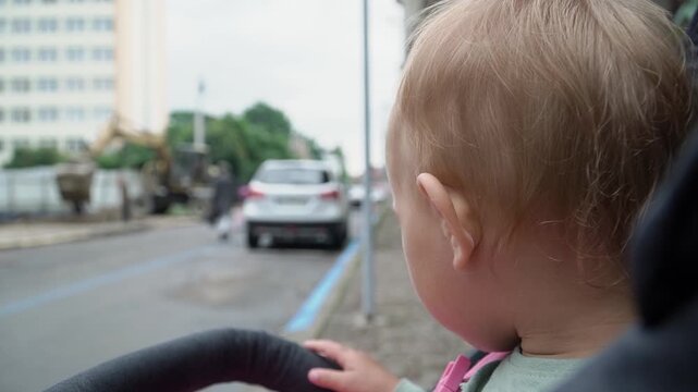 A baby with short hair green sweater sits stroller and watches the work of a construction excavator on the other side of the road, turns to the camera and looks surprise, on a cloudy summer day.