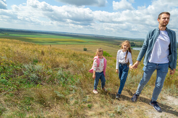 Fototapeta premium Father and daughter stand on top of hill and enjoy beautiful scenery of green hills under blue cloudy sky.