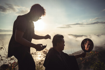 Silhouette of a Barber Cutting Hair Outdoors at Sunset with a Reflection in a Mirror Amidst a...