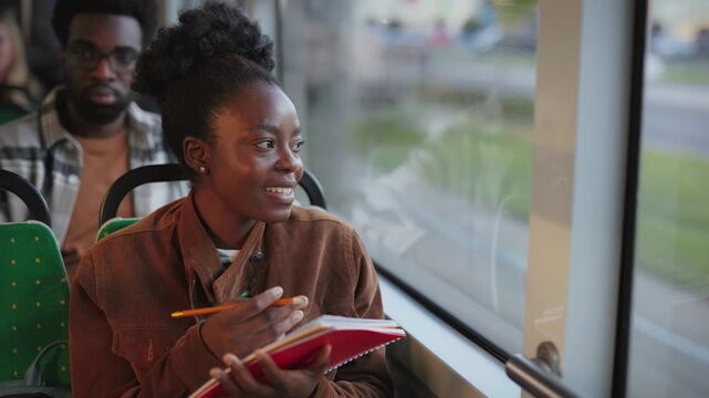 African American cheerful woman holding red notebook and writing with pencil while riding tram. Female student making notes smiling near window during travel. Thinking while looking outside.