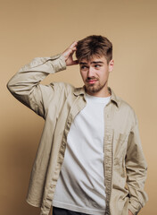 portrait of young man appears to be in a state of confusion, scratching his head on beige background