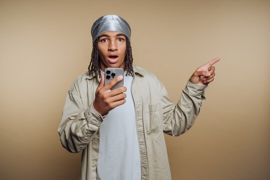 portrait of young man in a silver head wrap shows surprise while holding a smartphone on beige background