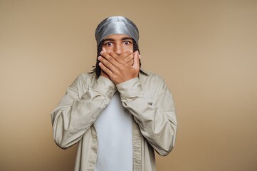 Fototapeta premium portrait of young startled man covers his mouth with hands, illustrating surprise and incredulity on beige background