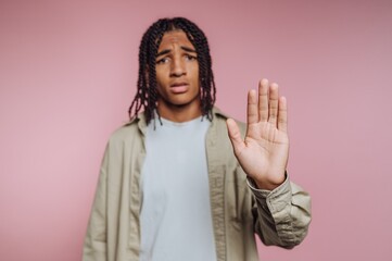 portrait of young man raises his hand in a gesture of denial or pause, expressing disbelief on pink background