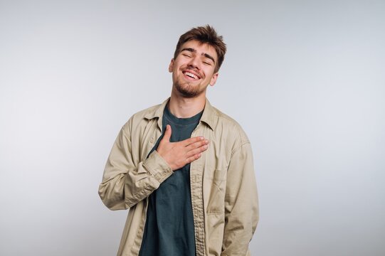 portrait of young man stands with his hand on his chest, wearing a warm smile that conveys gratitude and sincerity on white background