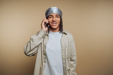 portrait of young man in a silver head wrap talks on his smartphone, showcasing communication on beige background