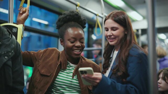 African American woman and Caucasian woman standing together in tram laughing while looking at smartphone screen. Friends enjoying shared moment during evening ride with other passengers around.