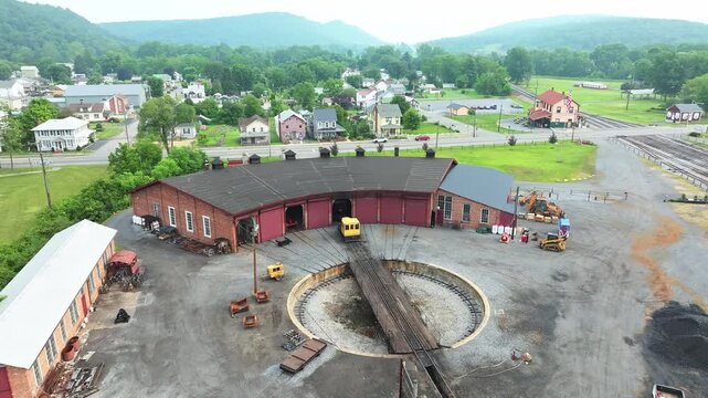 Visit a charming train yard surrounded by mountains. Discover red wooden roundhouse and buildings, rail tracks, and lush greenery. Experience the quiet beauty of this historical site on a sunny day