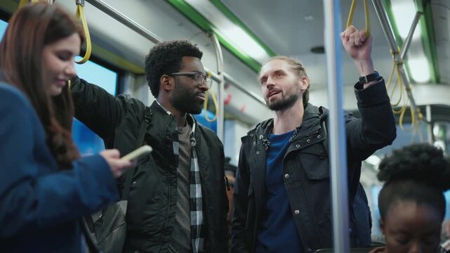 African American man with glasses standing besides bearded man. Talking inside metro. Both holding rails while smiling. Other passengers using phones and sitting nearby during ride.