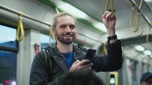 Handsome Caucasian man with long hair tied in bun standing in tram holding strap and smiling at camera. Male wearing dark jacket using smartphone with one hand during evening ride on public transport.
