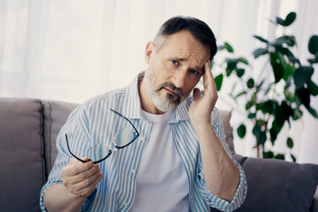 Mature man holding glasses and expressing stress while seated in a bright living room with indoor plants around