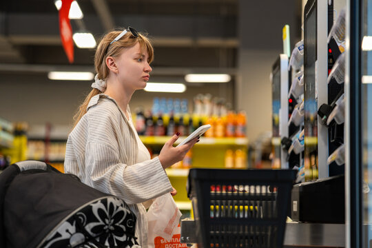 Young woman standing at self-checkout with shopping basket and phone in hand. Modern retail technology and payment process during grocery shopping.