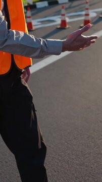 Person in an orange safety vest and black pants using hand signals to direct traffic or vehicles in a parking lot, working to guide movement and ensure safety in a striped outdoor area with cones
