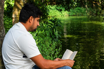 Fototapeta premium Peaceful moment as a young man enjoys reading a book by the serene lake, surrounded by lush greenery, embracing quiet reflection and intellectual stimulation in nature's embrace