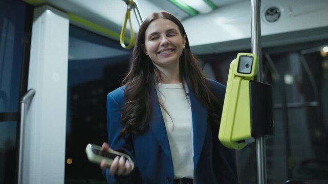 Pretty Caucasian woman with long hair holding smartphone and validating ticket on public transport. Female smiling while using contactless payment system inside modern city bus at night.