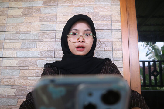 Young Asian woman in hijab and glasses talking into a smartphone camera for a video call or vlog. She has an engaged and slightly surprised expression, shot from a low angle.
