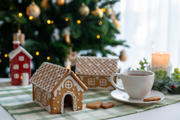 Handcrafted gingerbread houses with decorative icing and heart cookies beside white cup on festive table with Christmas tree background. Concept of homemade holiday treats, seasonal baking activities.