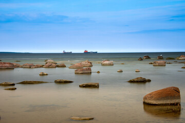 A serene shallow shoreline with scattered rocks, calm reflective water, and a soft blue sky...