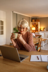 Happy elderly woman enjoying coffee while video calling therapist on laptop during online therapy...