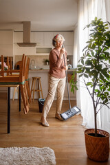 Senior woman listening to music on headphones dancing and cleaning parquet floor with mop and bucket in dining room