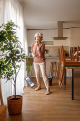 Mature woman in headphones holding mop stick as microphone and singing joyfully while mopping floor in kitchen