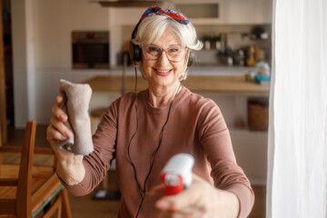 Portrait of happy old woman in headphones listening to music holding spray bottle and cleaning cloth at home