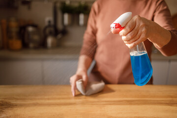 Midsection of mature woman's hand spraying disinfectant on wooden table while cleaning and dusting in kitchen at home