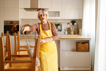 Portrait of cheerful mature woman wearing apron singing while dusting dining room chairs with duster at home