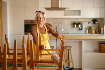 Mature woman wearing apron cleaning dust and dirt from wooden dining chairs with duster in modern kitchen