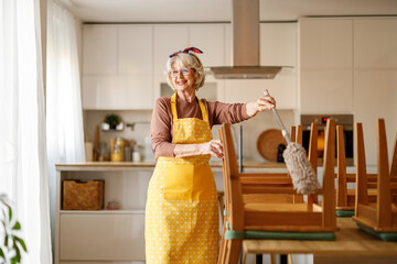 Smiling mature woman dusting wooden dining chairs with duster while tidying up modern kitchen at home