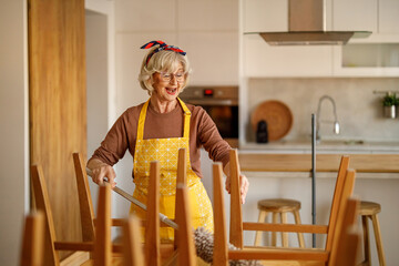 Cheerful senior woman in apron singing joyfully while dusting wooden dining chairs with duster in modern kitchen