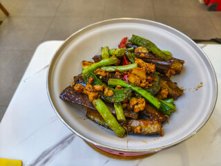 Delicious eggplant stir-fry with minced chicken and green chilies, served in a vibrant red bowl at a bustling restaurant.