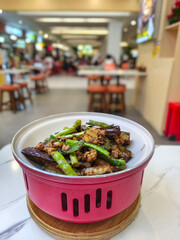Delicious eggplant stir-fry with minced chicken and green chilies, served in a vibrant red bowl at a bustling restaurant.