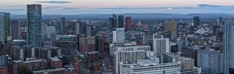 Telephoto panorama of Manchester skyline at dusk with a full Moon rising above the Pennine hills.