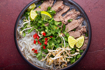 Pho Bo Vietnamese soup, bowl of fresh asian soup with rice noodle, beef meat, herbs, soy sprouts and chili on red background, top view close up. Asian cuisine, comfort food.