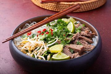 Pho Bo Vietnamese soup, bowl of fresh asian soup with rice noodle, beef meat, herbs, soy sprouts and chili on red background close up. Asian cuisine, comfort food.