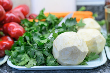 Peeled celery roots with fresh celery leaves arranged on a tray beside red bell peppers and carrots in a bright kitchen, showing preparation for homemade pickled vegetables.