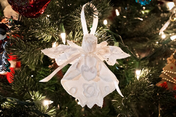 Close-up color photo of a handmade white fabric angel on a green plastic tree with lights and soft...