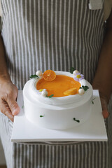 Close-up of a person holding a homemade orange cake decorated with mandarin slices, pearls, and flowers,Minimal style.