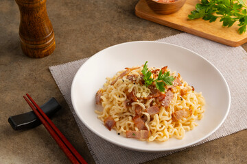 Instant noodle carbonara sauce topped with crispy bacon and parmesan cheese in white plate.