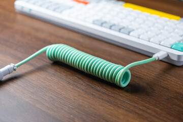 Mint green coiled braided USB cable connected to a modern mechanical keyboard on a wooden desk, enhancing desktop aesthetics for work or gaming