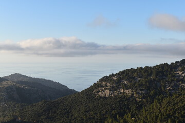 Dramatic landscape of the Serra de Tramuntana in Mallorca, featuring lush green hills, rugged peaks, and the Mediterranean Sea under a serene blue sky with scattered clouds
