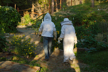 Two Beekeepers in full White Protective gear Suits Walk Through a Garden Path Among Flowers. Surrounded by blooming flowers and lush greenery. The scene conveys outdoor work, safety, and harmony with 
