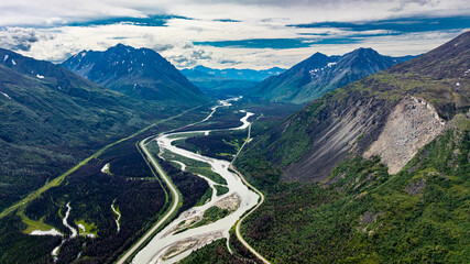 Fototapeta premium Winding river in Alaskan mountain valley aerial view. Aerial view of a winding river between Alaskan mountains with lush green vegetation and cloudy sky.