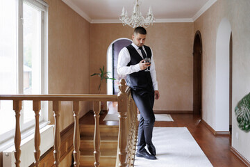 Man in formal attire holding drink while standing at staircase in elegant hallway