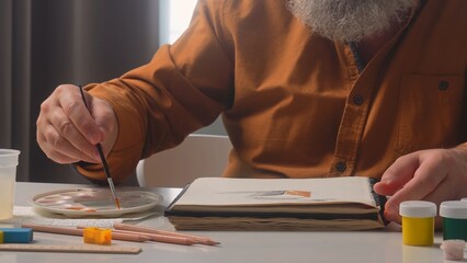 An elderly male artist attentively painting at a home studio table, surrounded by art supplies. © kinomaster