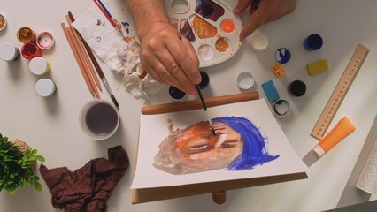 An older male artist paints a portrait on a canvas. His art supplies and painting equipment...